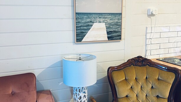 Une pièce avec un plancher en bois, un mur blanc orné d’un tableau représentant un quai, un fauteuil marron, une chaise rouge et une lampe posée sur une table d’appoint en bois.