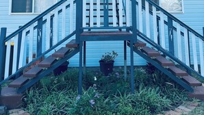 Un bâtiment bleu avec un escalier central menant à une porte, le tout entouré d’un jardin où poussent différentes espèces de plantes.