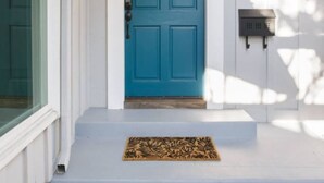 Une porte bleue avec une petite fenêtre, une boîte aux lettres et un paillasson sur un porche blanc.