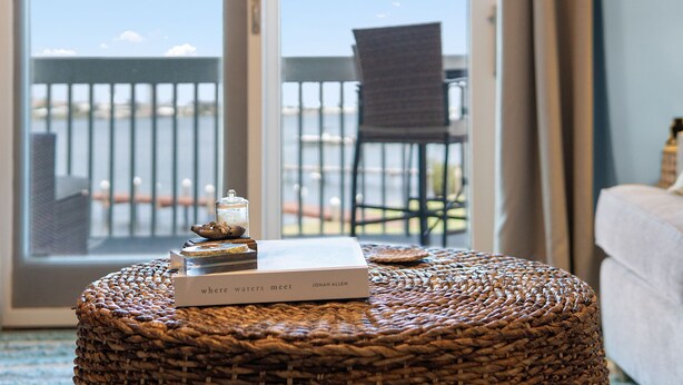 Una habitación con un reposapiés de mimbre, un libro titulado 'Where Waters Meet' de Jonah Allen, y vistas a un balcón con una silla.