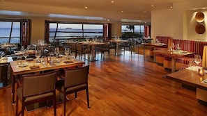 Ein Restaurant mit Holzboden, mehreren Esstischen und Stühlen. Große Fenster bieten Blick auf den Ozean.