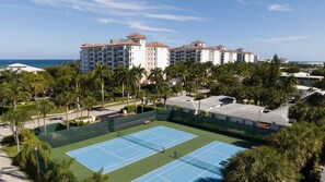 Un campo da tennis circondato da palme e un hotel con diversi piani e balconi.