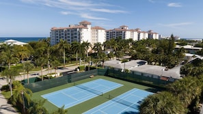 A tennis court surrounded by palm trees and a hotel complex with multiple buildings.