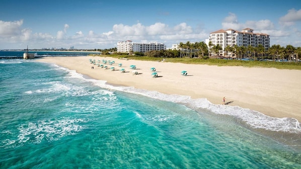 A beach with clear turquoise waters, a row of beach umbrellas, and a hotel with multiple stories and balconies overlooking the beach.