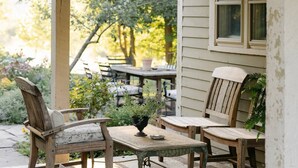 A covered outdoor seating area with a table and chairs, a potted plant, and a window overlooking a garden. - Hudson Pond · Garden Queen Suite · GF Breakfast (Poughkeepsie)