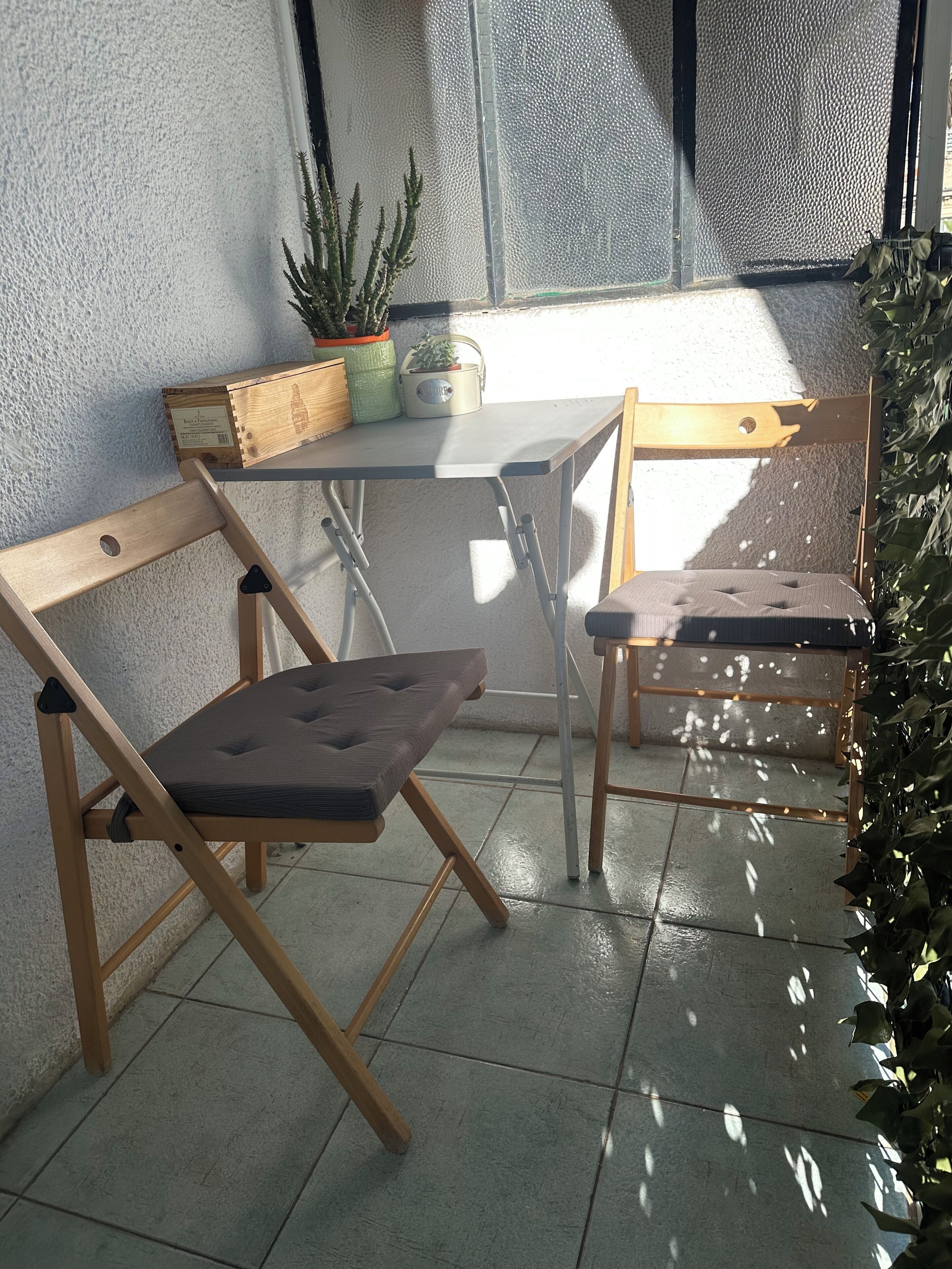 Un pequeño espacio exterior con una mesa y dos sillas, una planta en maceta y una ventana con vistas.