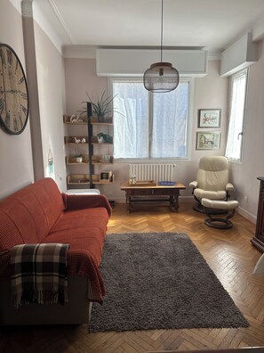 A living room with a sofa, a chair, a rug, a radiator, a window with curtains, a hanging light fixture, and a clock on the wall. - Bright apartment in the heart of San Remo (Sanremo)