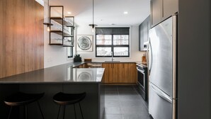 A modern kitchen with a central island, stainless steel appliances, wooden cabinets, and a large window. - Le Contemporain by Gestion ELITE (Mont-Tremblant)