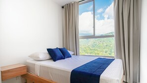 A bedroom with a wooden bed, white bedding, blue pillows, and a view of a green landscape through a window. - MonteAzul apto con vista a la Cordillera (Armenia)