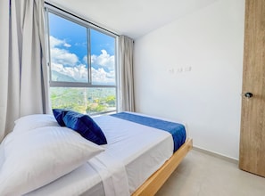 Un dormitorio con cama, ventana con cortinas, vista a zonas verdes y una puerta de madera.