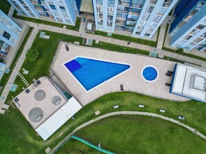 Vue aérienne d’un complexe immobilier moderne, avec une grande piscine bleue, une fontaine circulaire et un petit bâtiment doté d’une fenêtre circulaire.