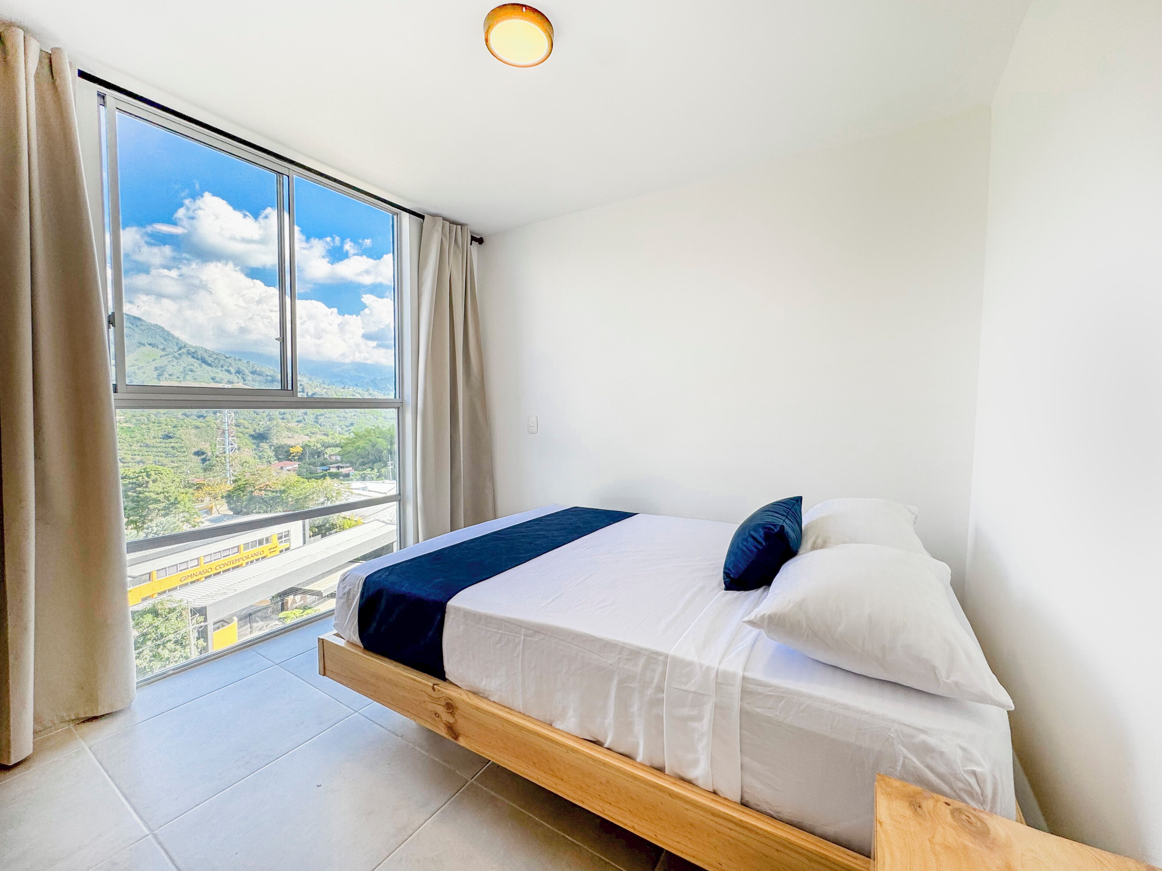 Una habitación con una cama, una ventana con cortinas, una lámpara en el techo y vistas a la vegetación y un tren.