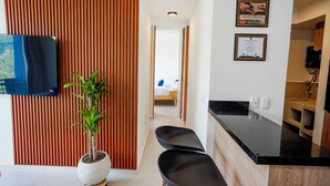 Et moderne hotelværelse med fjernsyn, en potteplante, en træafskærmning, et tekøkken og en gang der fører til et soveværelse.