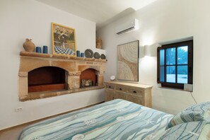 A bedroom with a fireplace, a bed with patterned bedding, a dresser, and a window with a view. - Exclusive Villa Qupi in the center of Lindos separate staff accommodation (Lindos)