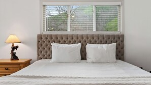 A bedroom with a tufted headboard, two white pillows, a bedside lamp, and a window with blinds. - The Hangout Cabin by Avantstay Hot Tub + Game Room + Firepit + Near Gatlinburg + Dollywood (Gatlinburg)