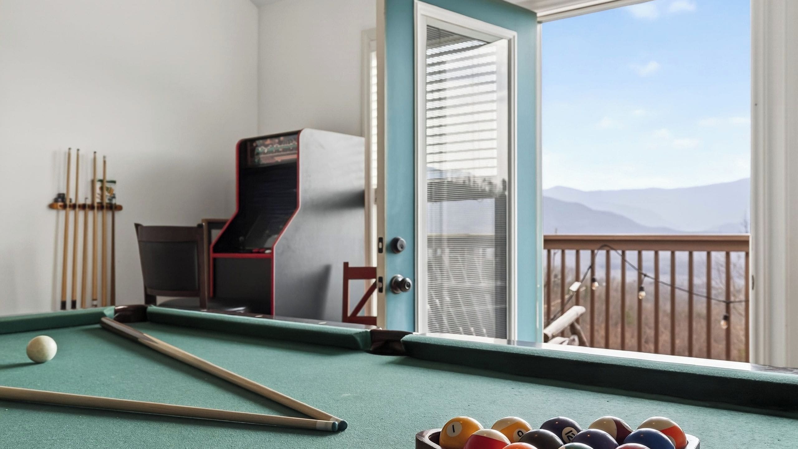 A pool table with balls, a pool cue, and a billiards rack in a room with a view of mountains through a door.