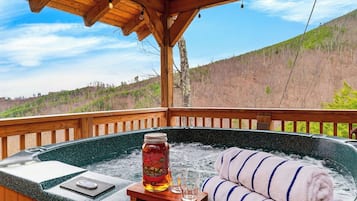 A hot tub with a bottle of liquor and a towel on a wooden deck with a view of a forested hillside.
