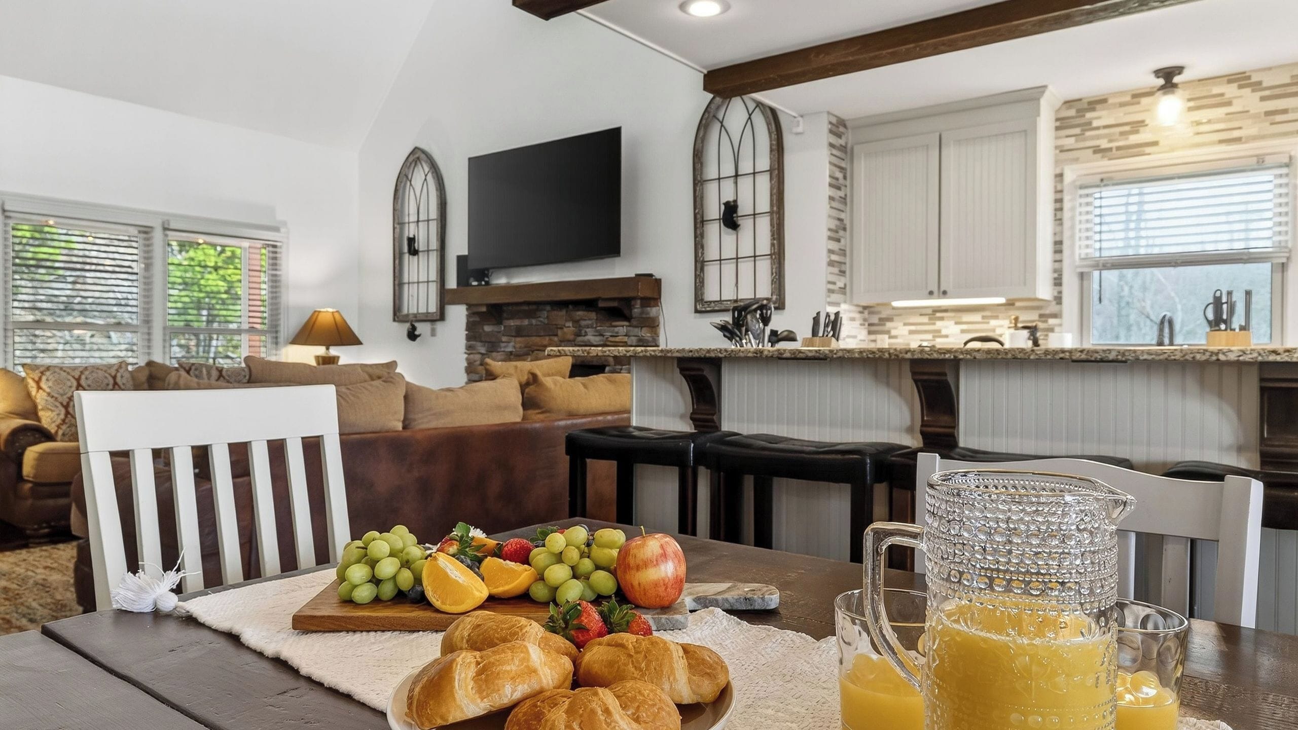Une cuisine moderne avec un mur recouvert de pierres, un téléviseur à écran plat et un coin repas où une table est dressée avec des fruits et des pâtisseries.