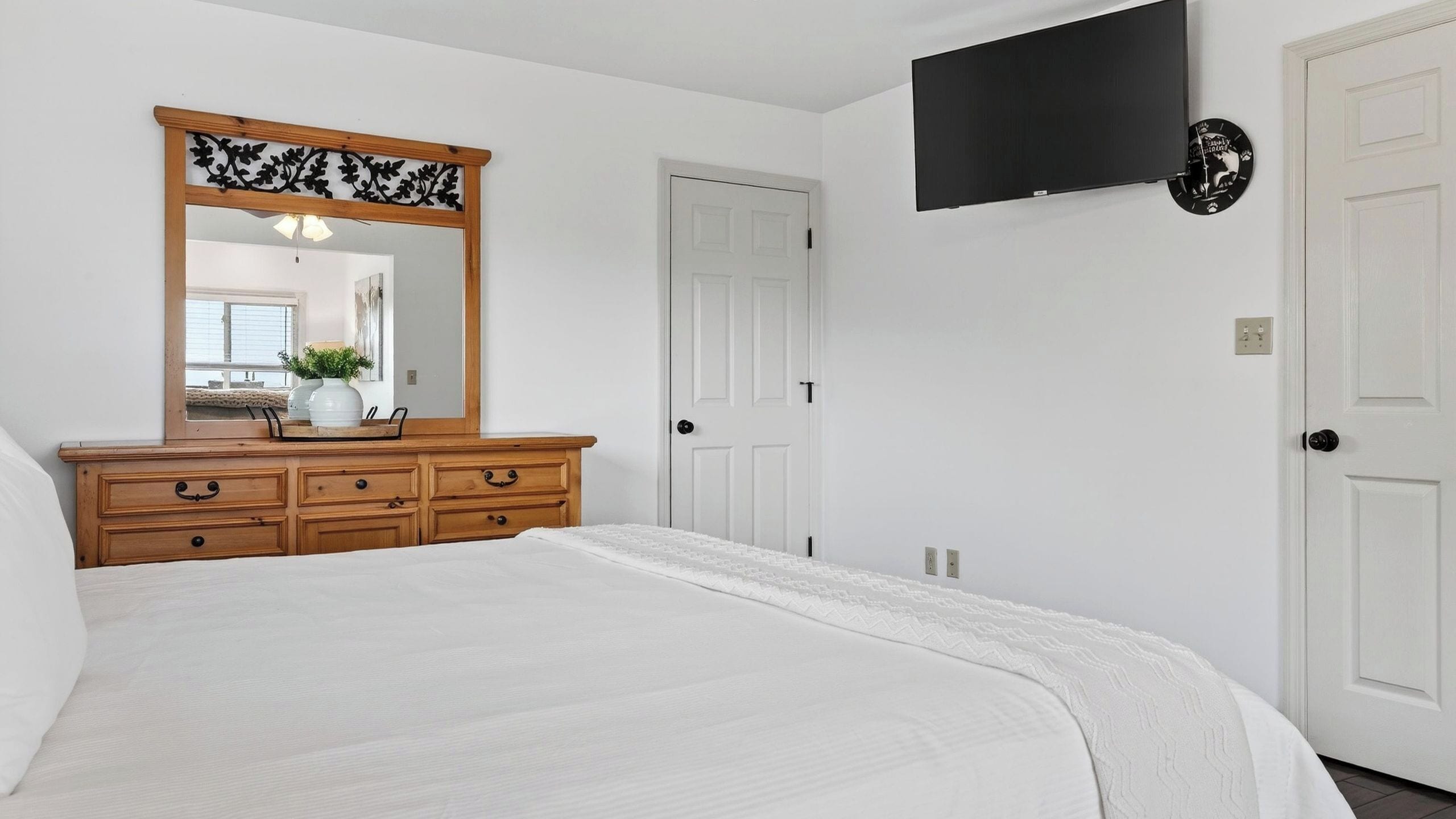 A bedroom with a bed, wooden dresser, mirror, white walls, and a mounted TV.