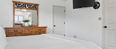 A bedroom with a bed, wooden dresser, mirror, white walls, and a mounted TV.
