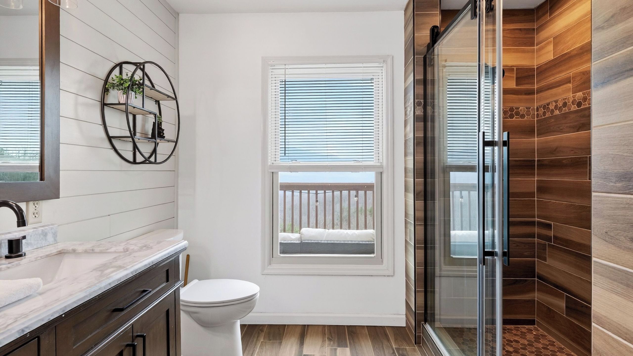 A bathroom with a toilet, a sink, a mirror, a window with blinds, and a shower area with wooden walls.