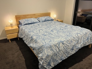 A bed with a leaf-patterned bedspread, two bedside tables with lamps, and a wooden headboard. - Casa Acta - Reel Road (Waihi Beach)