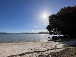 A beach with a clear sky, calm water, and distant mountains. - Casa Acta - Reel Road (Waihi Beach)