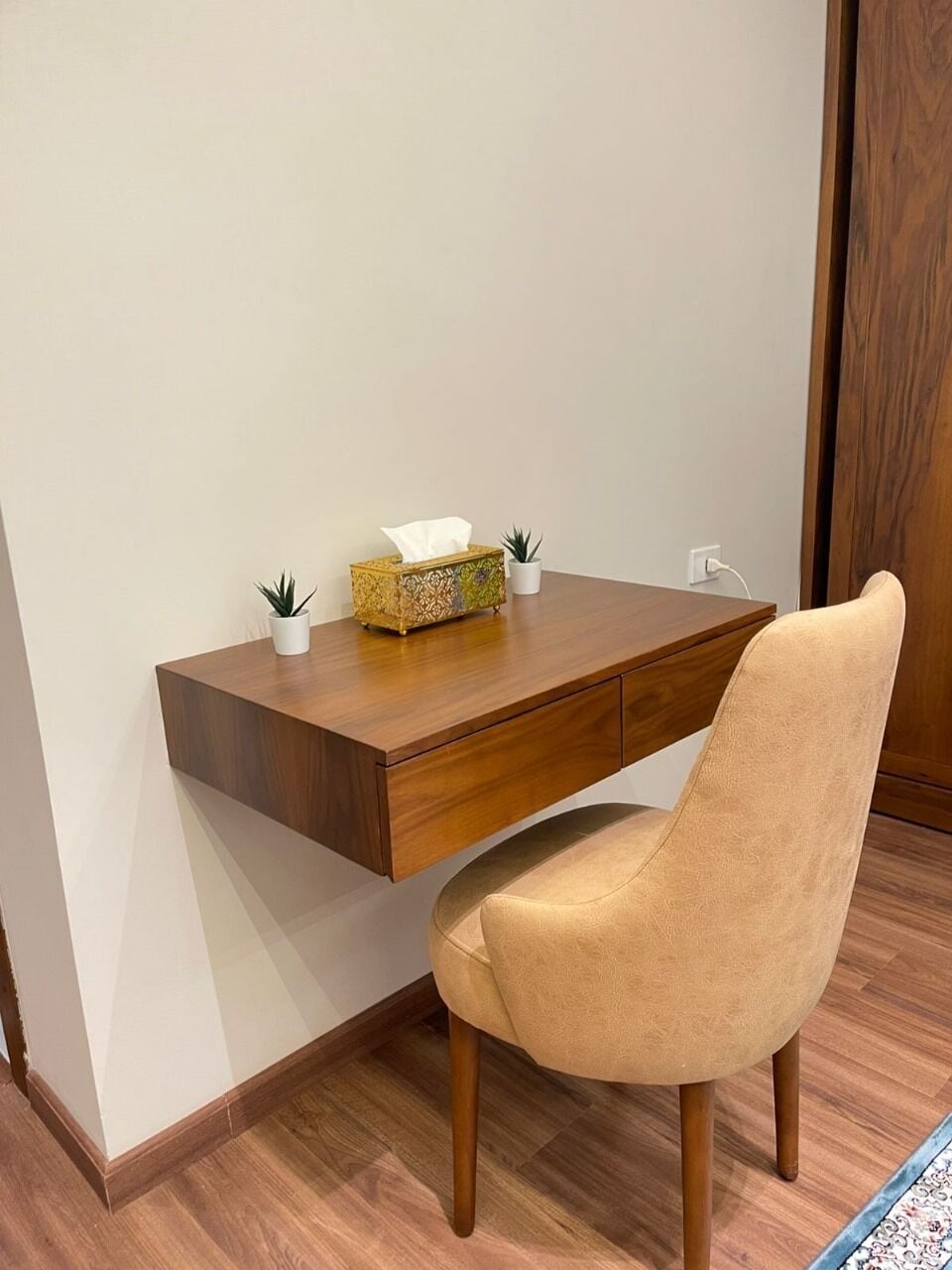 Uma escrivaninha de madeira com uma cadeira estofada, uma caixa de lenços de papel e duas pequenas plantas em vasos.