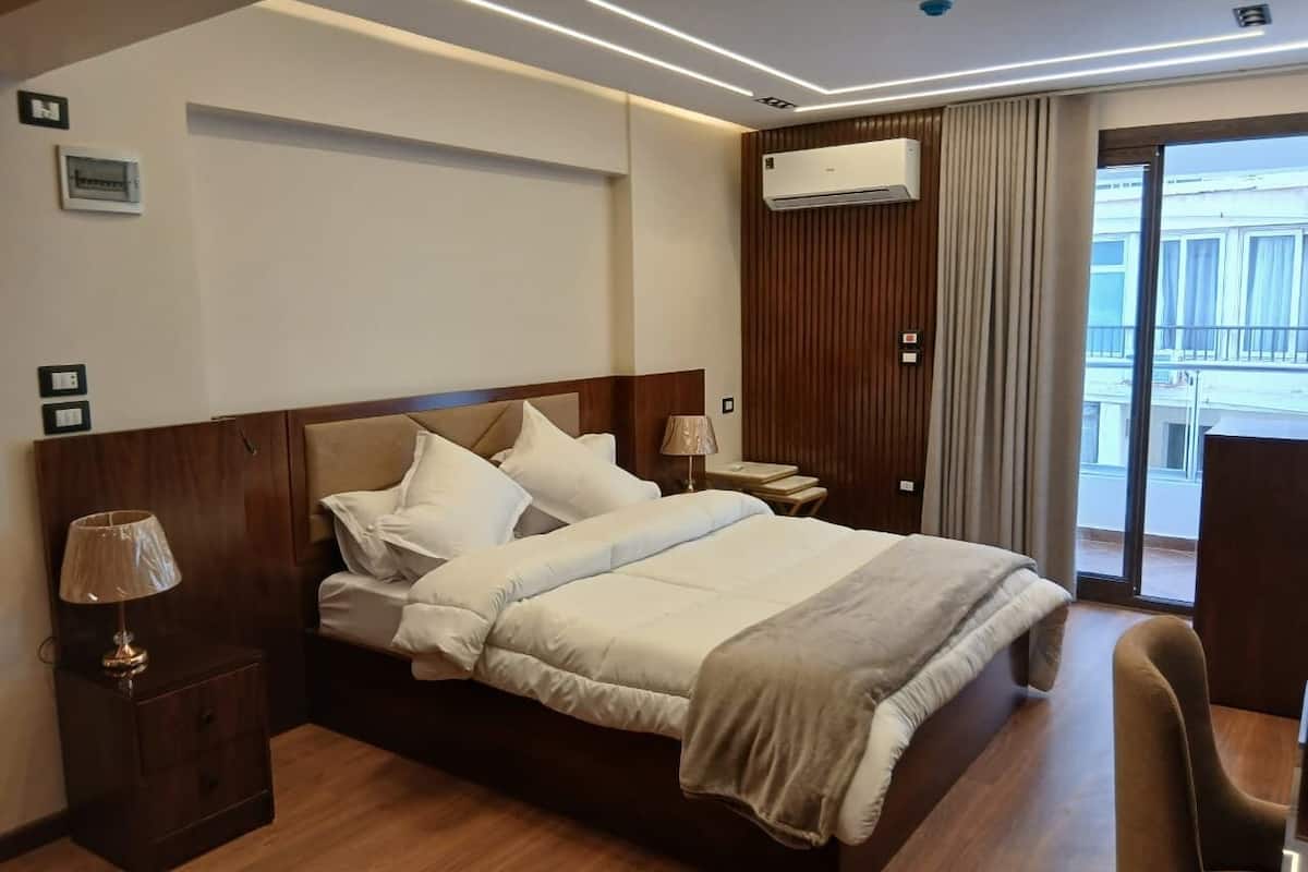 A hotel room with a bed, bedside tables, a lamp, an air conditioning unit, and a view of the outside.