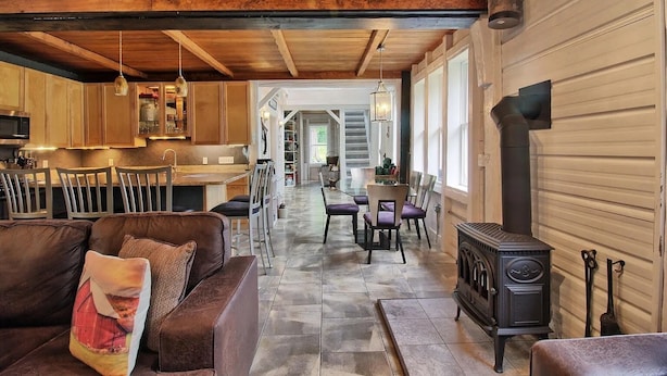 A rustic interior with a wooden ceiling, a dining area, a kitchen with wooden cabinets, and a wood-burning stove. - The Old Maine Barn - Near Acadia & Bar Harbor (Ellsworth)
