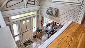 A loft-style room with a high ceiling, exposed wooden beams, a large wooden wheel, and a view of the lower level through a glass railing. - The Old Maine Barn - Near Acadia & Bar Harbor (Ellsworth)