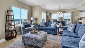 A living room with a view of the ocean, a blue sofa set, a wooden ladder shelf, and a chandelier. - Stunning corner unit in the heart of Pensacola beach with panoramic views. (Pensacola Beach)