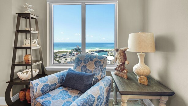 A room with a view of the beach, a chair with a patterned cushion, a lamp, and a shelf with decorative items. - Stunning corner unit in the heart of Pensacola beach with panoramic views. (Pensacola Beach)