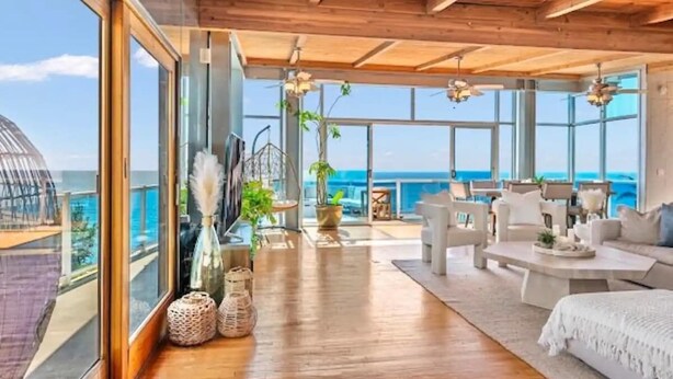 Una habitación amplia con techo de madera, ventanas grandes, un sofá blanco y vistas al océano.