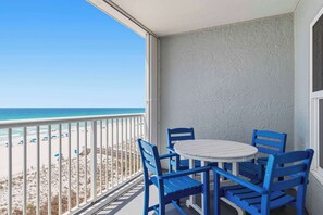 A balcony with a white table and blue chairs, overlooking a beach with umbrellas. - Spacious gulf front condo with beach access and seasonal beach service (Okaloosa Island)