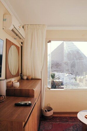 A hotel room with a wooden desk, a television, a large window with a view of a mountain, and a red patterned rug. - Pyramids View • places (محافظة الجيزة)