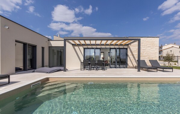 Un espace piscine extérieur moderne agrémenté d’une pergola, d’une table à manger avec des chaises et d’un coin salon avec des chaises longues.