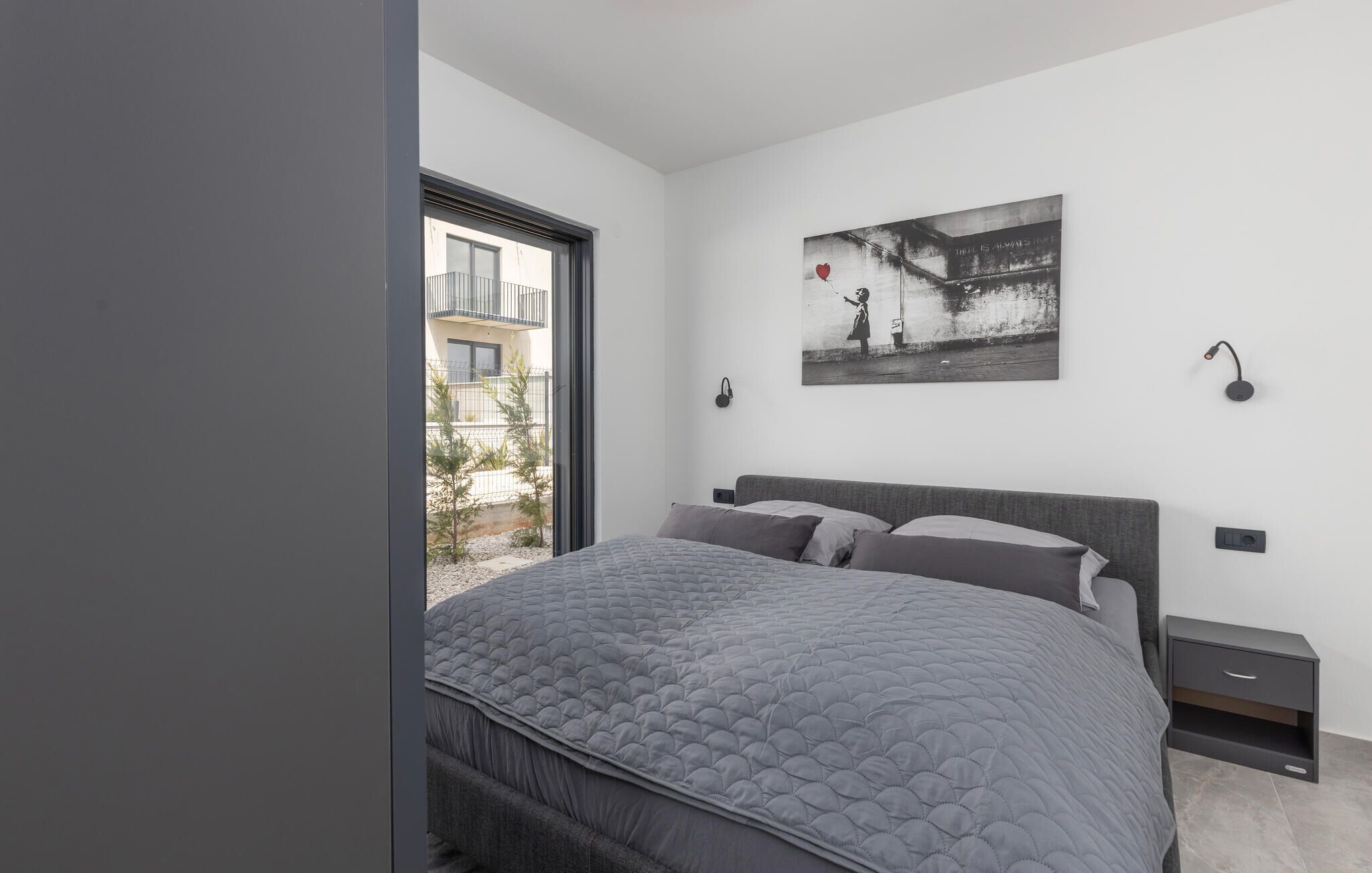Une chambre moderne avec un grand lit, un tableau au mur, une table de chevet et une vue sur l’extérieur.