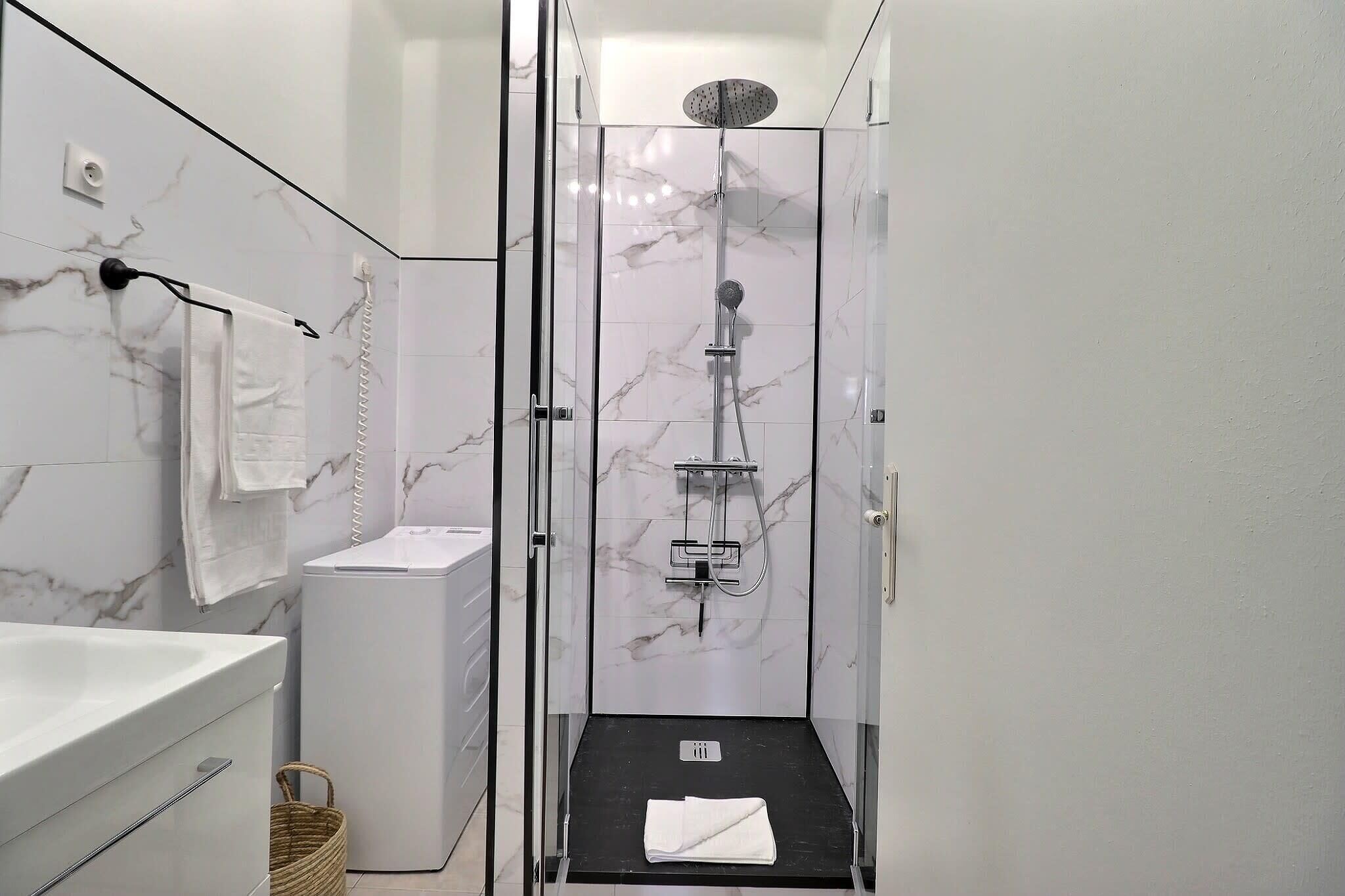 Un bany modern amb dutxa tancada per vidre, paret de marbre blanc, un lavabo blanc i un paviment negre.