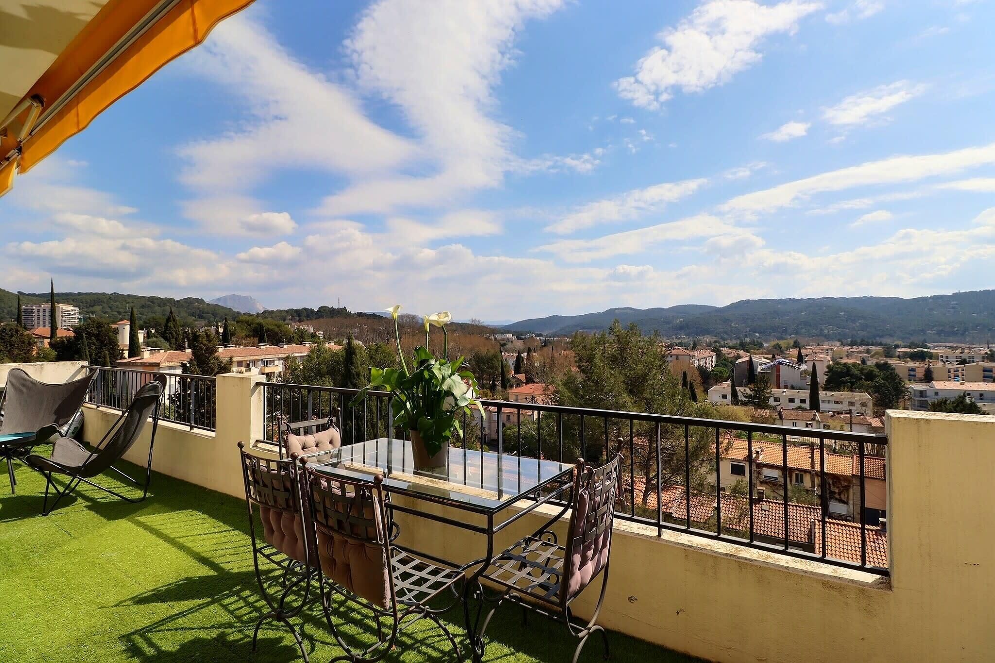 A balcony with a table and chairs, a potted plant, and a view of a cityscape with buildings and hills.