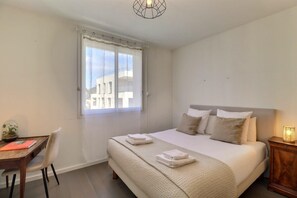 Ein Schlafzimmer mit Bett, Nachttisch, Lampe, Fenster und Schreibtisch mit Stuhl.