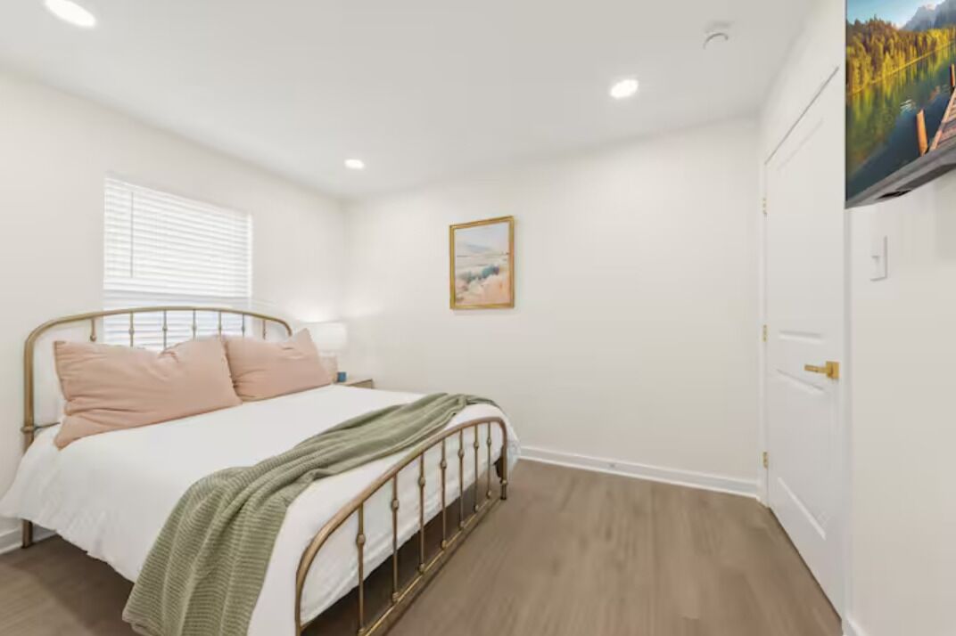 A bedroom with a bed, a window with blinds, a framed picture on the wall, and a door with a picture on it.