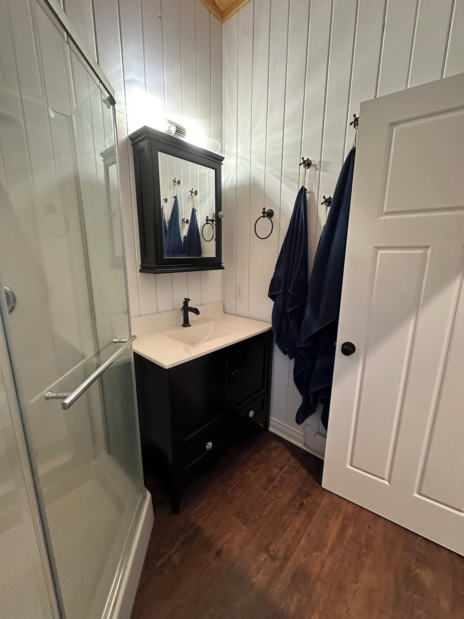 A bathroom with a glass shower enclosure, a sink with a mirror, and a door with towels hanging on hooks.