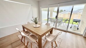 Un comedor con una mesa de madera, cuatro sillas, una planta en maceta y un puerta corrediza de vidrio que da a un patio exterior con una parrilla.