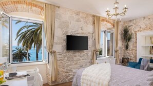 Een slaapkamer met een stenen muur, een bed met een beddeken met dessin, een televisie en uitzicht op palmbomen door een raam.