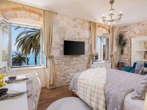 Ein Schlafzimmer mit einer Steinwand, einem Bett mit gemustertem Überwurf, einem Fernseher und Blick auf Palmen durch ein Fenster.