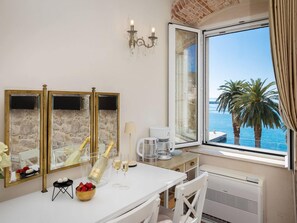 Ein Hotelzimmer mit großem Fenster, einem weißen Tisch mit Champagnerflasche, zwei Gläsern, einer Erdbeerschale, einer Kaffeemaschine und einem Spiegel mit steinartigen Verkleidungen.