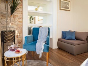 Ein Wohnzimmer mit blauem Sessel, braunem Sofa, einem kleinen runden Tisch mit Teekanne und einem Bücherregal im Hintergrund.