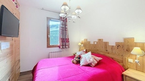 Une chambre à coucher avec une tête de lit en bois, un lit recouvert d’une couvre-lit rose vif, un téléviseur fixé au mur, une fenêtre avec des rideaux et un lustre.