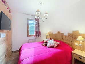 A bedroom with a wooden headboard, a bed with a bright pink bedspread, a television mounted on the wall, a window with curtains, and a chandelier. - 200m from resort center, 3rd floor, swimming pool, balcony, tv, ski locker, 40m², Le Corbier (Le Corbier)